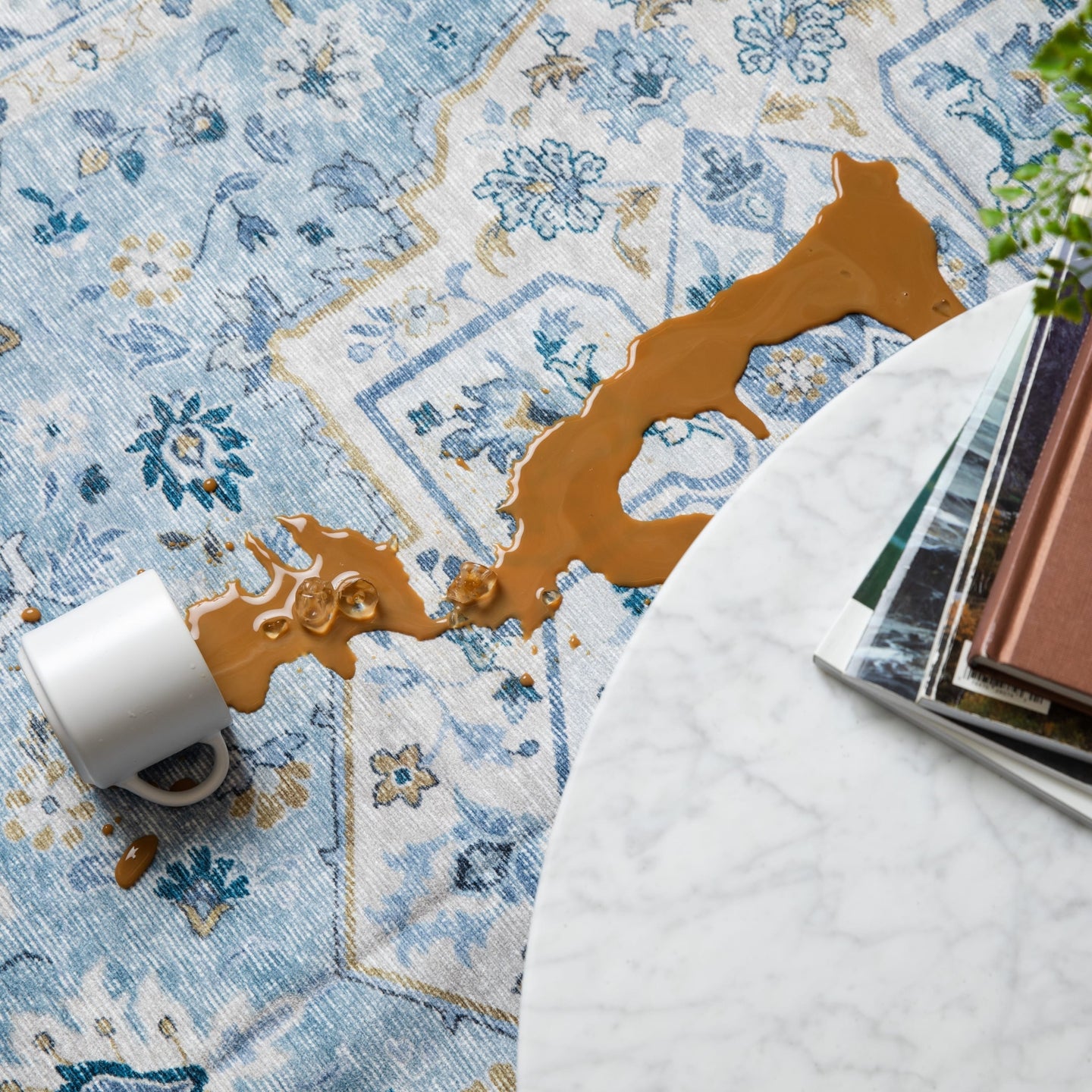 Spilled coffee on a blue patterned rug beside a white marble table with books.